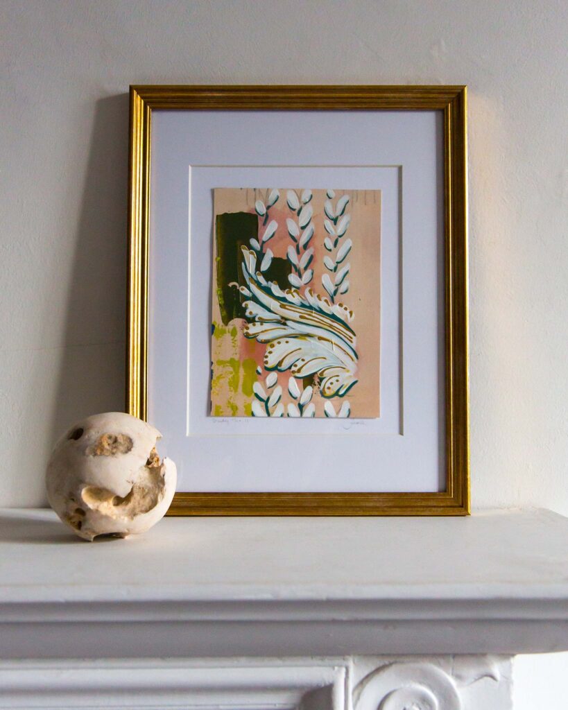 Framed architectural painting, Forma Coloris Lux Mentis — Study No. 11 by Teale Hatheway, displayed on a white mantel in a gold frame with a double mat. The artwork pairs a bold ornamental scroll with vertical leaf elements, highlighted by shimmering gilded details, accompanied by a textured stone sphere.