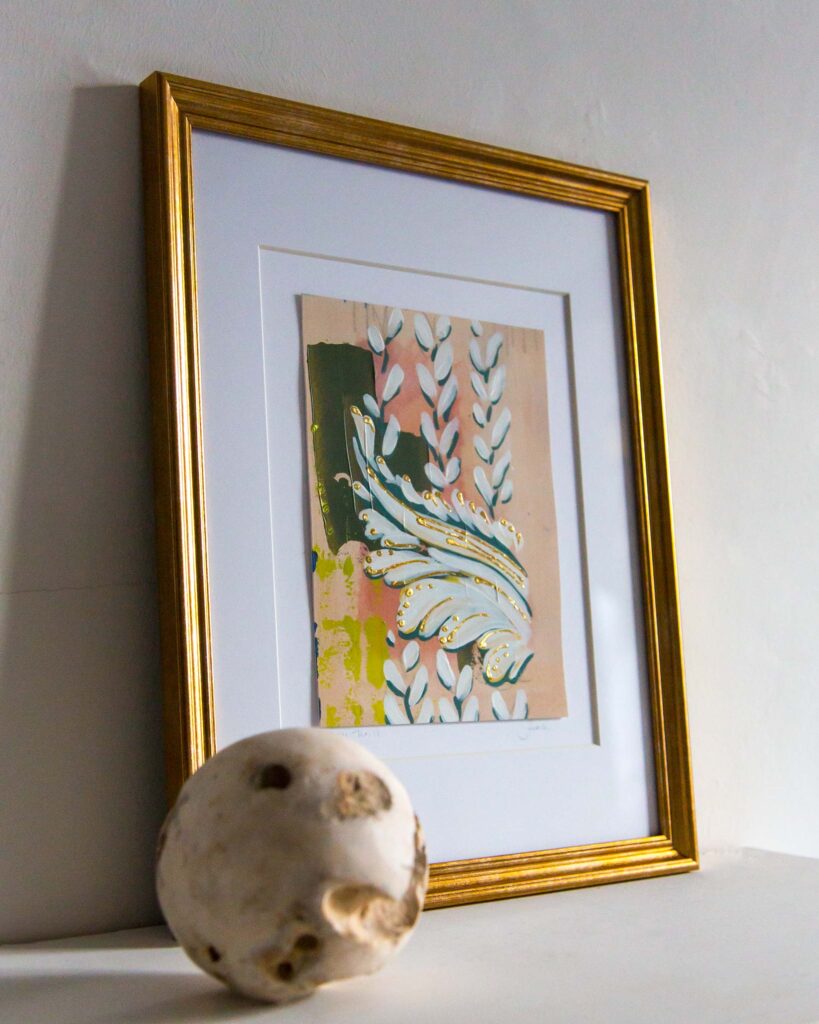 Angled view of Teale Hatheway’s Forma Coloris Lux Mentis — Study No. 11 framed in gold with a double mat, resting on a white mantel. The artwork features a sweeping ornamental flourish layered over vertical leaf motifs, with hand-applied gilding and abstract blocks of green, peach, and yellow. A sculptural stone sphere sits in the foreground.