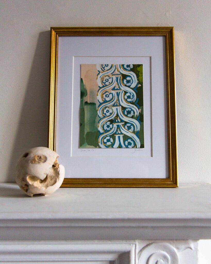 Framed architectural painting, Forma Coloris Lux Mentis — Study No. 13 by Teale Hatheway, displayed on a white mantel in a gold frame with a double mat. The artwork features a vertical ornamental column of scrolling blue-and-white motifs with gilded accents, set against layered tones of green, peach, and yellow. A sculptural stone sphere sits nearby.