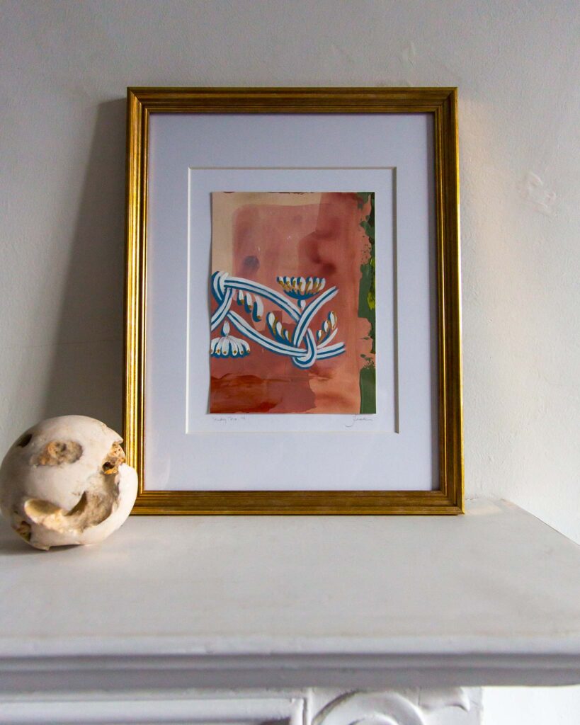 Framed architectural painting, Forma Coloris Lux Mentis — Study No. 14 by Teale Hatheway, displayed on a white mantel in a gold frame with a double mat. The artwork shows a braided blue ornamental line with petaled flourishes and gilded highlights over warm terracotta and olive tones, paired with a textured stone sphere.