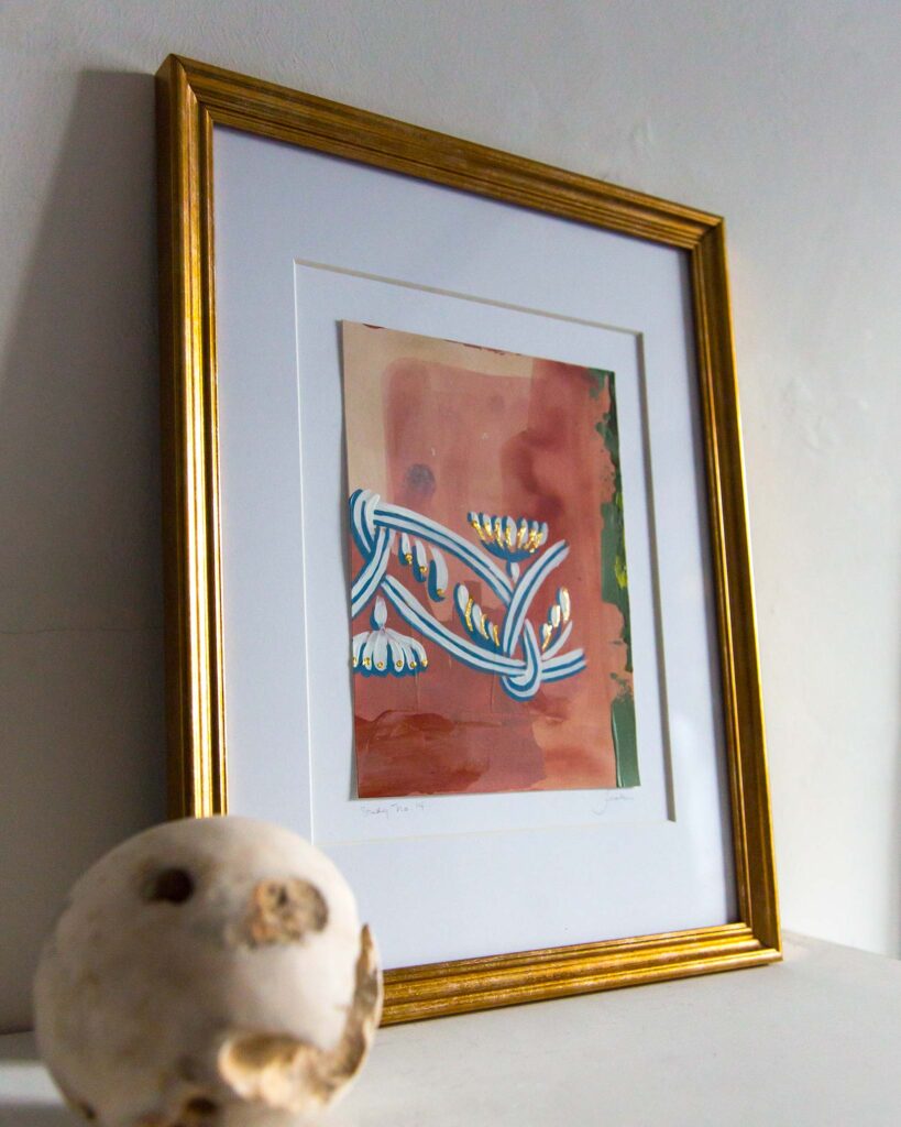 Angled view of Teale Hatheway’s Forma Coloris Lux Mentis — Study No. 14 framed in gold with a double mat, resting on a white mantel. The artwork features a blue-and-white braided ornamental motif with gilded accents against warm terracotta, peach, and green abstract layers, with a sculptural stone sphere in the foreground.