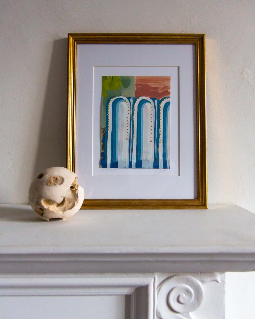 Framed architectural painting, Forma Coloris Lux Mentis — Study No. 15 by Teale Hatheway, displayed on a white mantel in a gold frame with a double mat. The artwork depicts three elongated blue arches highlighted with gold accents over layered green, peach, and terracotta tones, accompanied by a textured stone sphere.