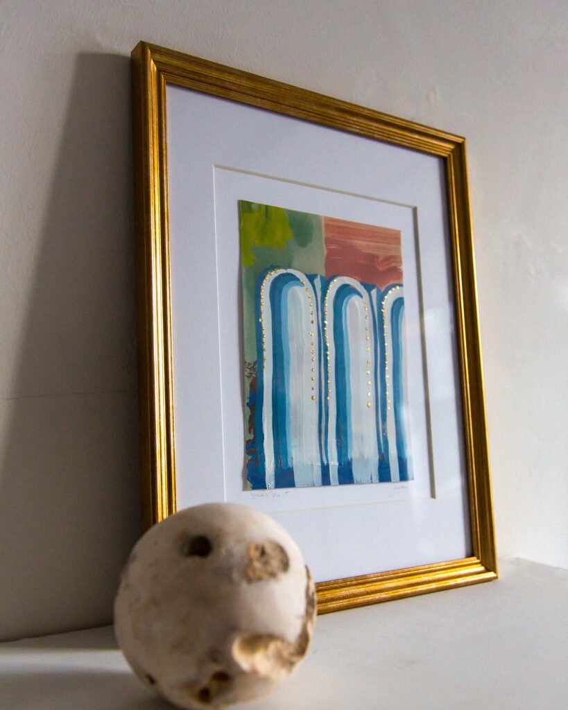 Angled view of Teale Hatheway’s Forma Coloris Lux Mentis — Study No. 15 framed in gold with a double mat, resting on a white mantel. The artwork features a trio of tall blue arches with gilded detailing set against abstract fields of green, peach, and terracotta, with a sculptural stone sphere in the foreground.