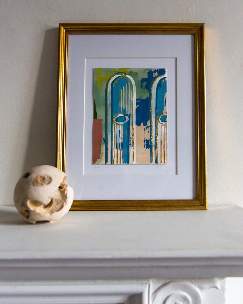 Framed architectural painting, Forma Coloris Lux Mentis — Study No. 16 by Teale Hatheway, displayed on a white mantel in a gold frame with a double mat. The artwork depicts vertical blue arches with gold-accented oval motifs over textured fields of green, pink, and ochre, accompanied by a textured stone sphere.