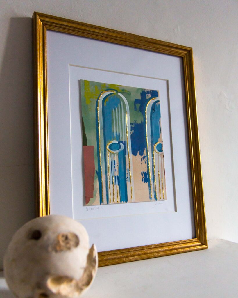 Angled view of Teale Hatheway’s Forma Coloris Lux Mentis — Study No. 16 framed in gold with a double mat, resting on a white mantel. The artwork features tall blue architectural arches with elongated oval details and hand-applied gilding, set against abstract blocks of green, pink, and ochre. A sculptural stone sphere sits in the foreground.