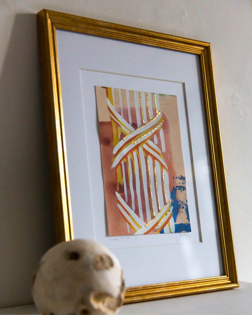 Angled view of Teale Hatheway’s Forma Coloris Lux Mentis — Study No. 2 framed in gold with a double mat, resting on a white mantel. The photograph highlights the dimensional gilding and layered architectural forms, with a textured stone sphere in the foreground.