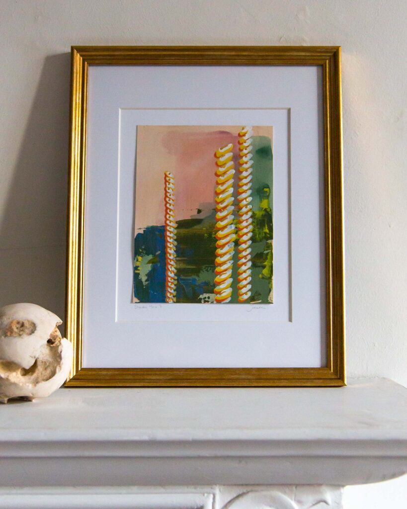 Framed architectural study, Forma Coloris Lux Mentis — Study No. 7 by Teale Hatheway, displayed on a white mantel in a gold frame with a double mat. The artwork features vertical, rope-like ornamental forms with gilded details against layered tones of pink, green, and deep blue, accompanied by a sculptural stone sphere.
