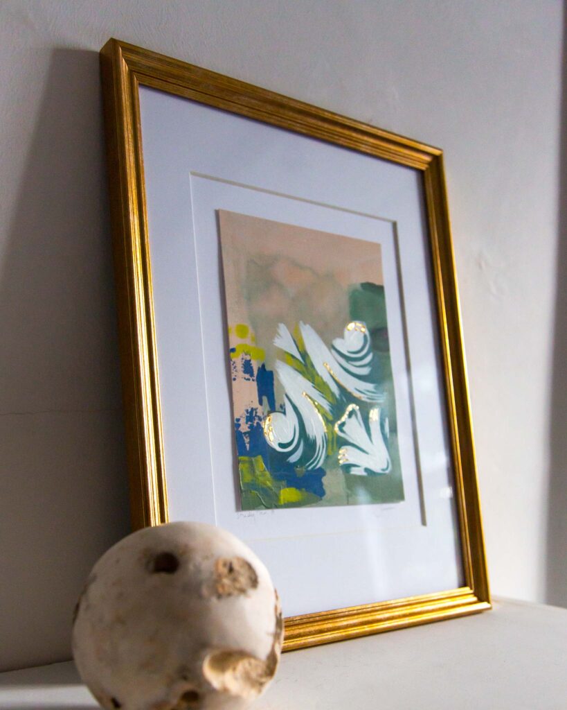 Angled view of Teale Hatheway’s Forma Coloris Lux Mentis — Study No. 8 framed in gold with a double mat, resting on a white mantel. The photograph highlights the gilded, swirling architectural motifs and the textured color fields behind them, with a stone sphere in the foreground.